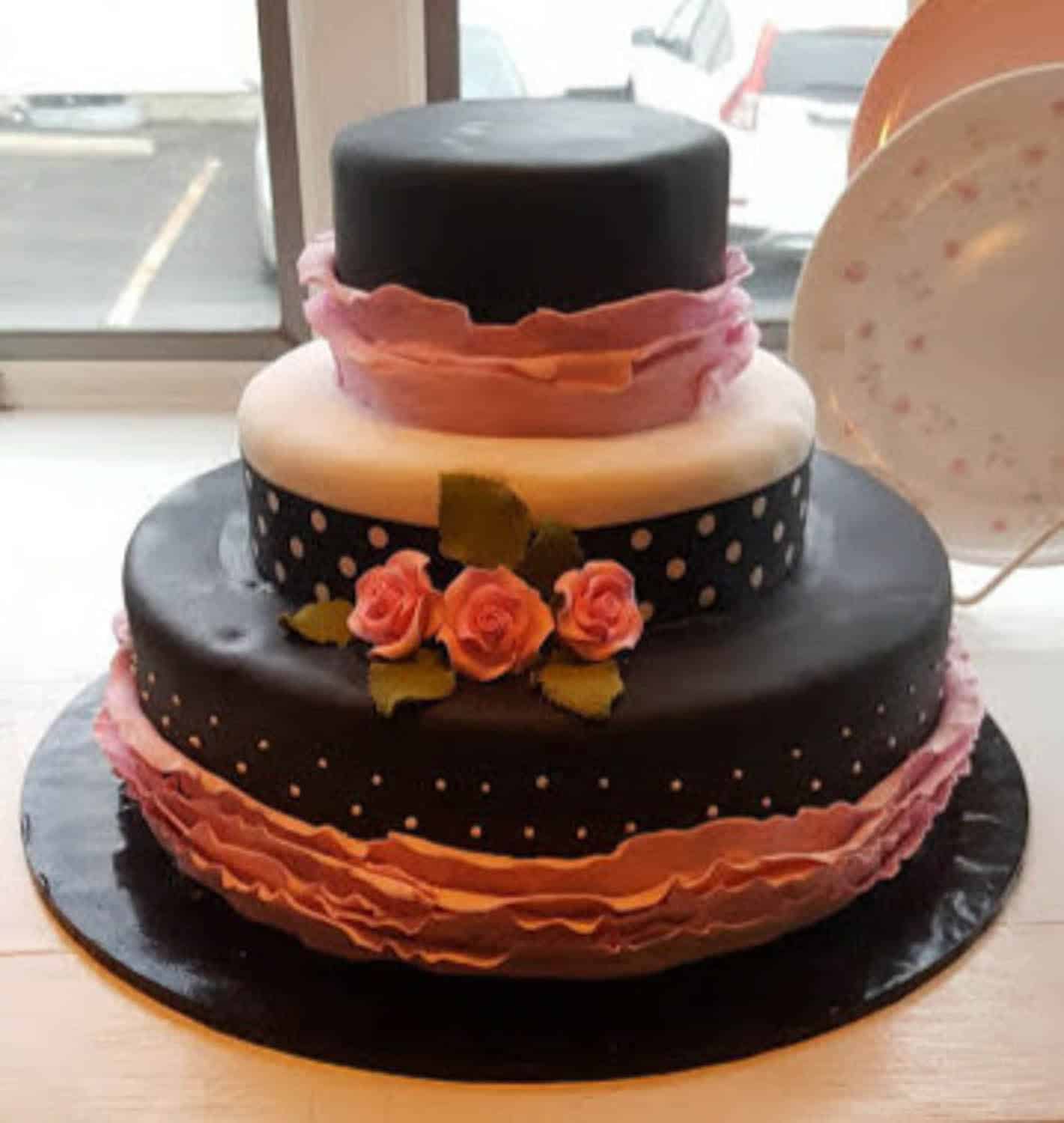 Three-tiered cake decorated with black and white fondant, pink ruffle accents, polka dots, and pink rose decorations, displayed on a black cake board near a window.