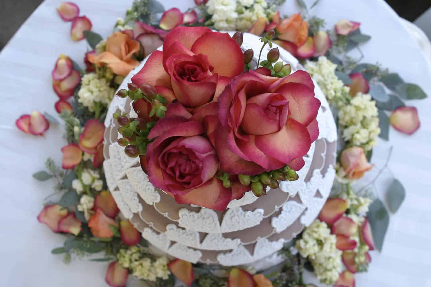 Round white tiered cake with intricate icing, topped with large pink roses, surrounded by flower petals and greenery on a white tablecloth.