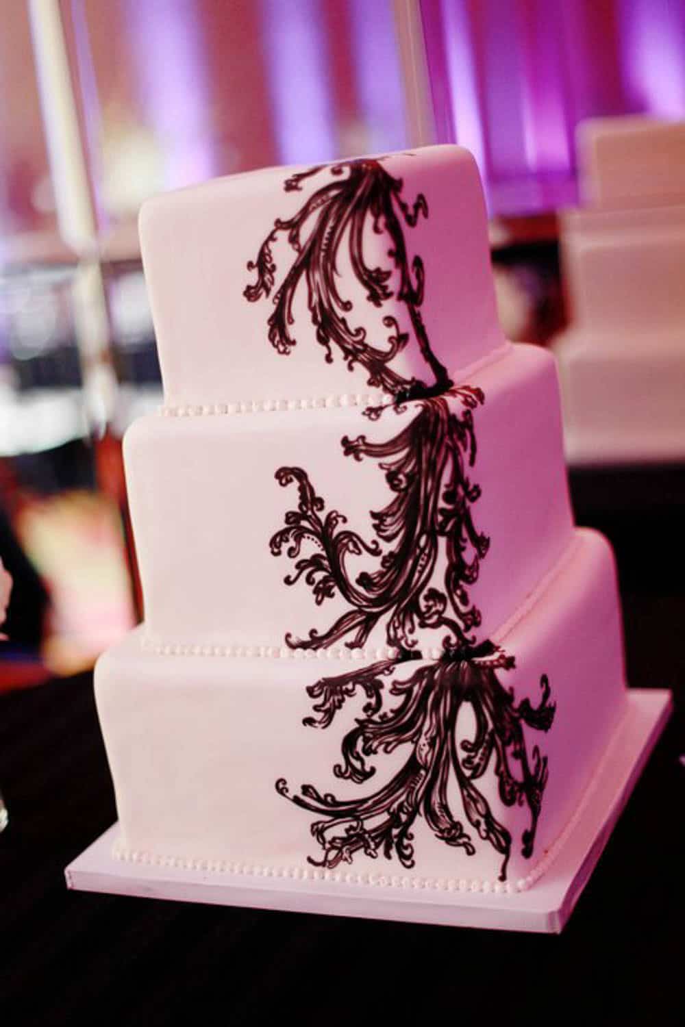 Three-tiered white cake with black ornate vine-like icing decoration cascading diagonally across each layer, set on a white square base.