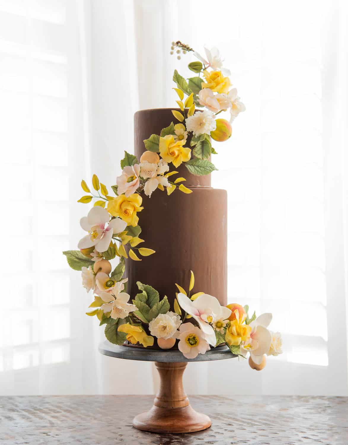 Two-tiered chocolate cake decorated with yellow, white, and peach sugar flowers, displayed on a wooden cake stand in front of a bright window.