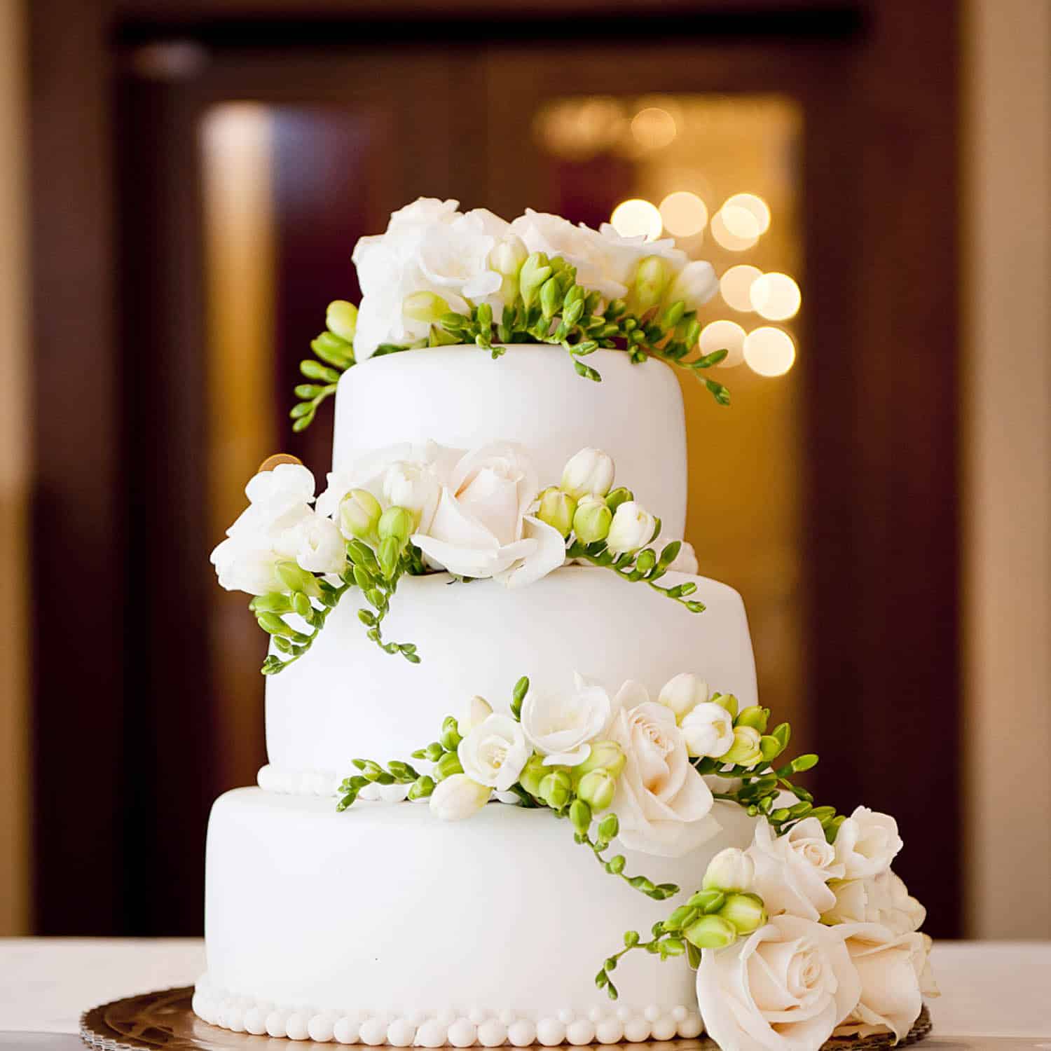 Three-tier white cake with white roses, green buds, and pearl-like edging at each tier base, set before blurred double doors with lights.