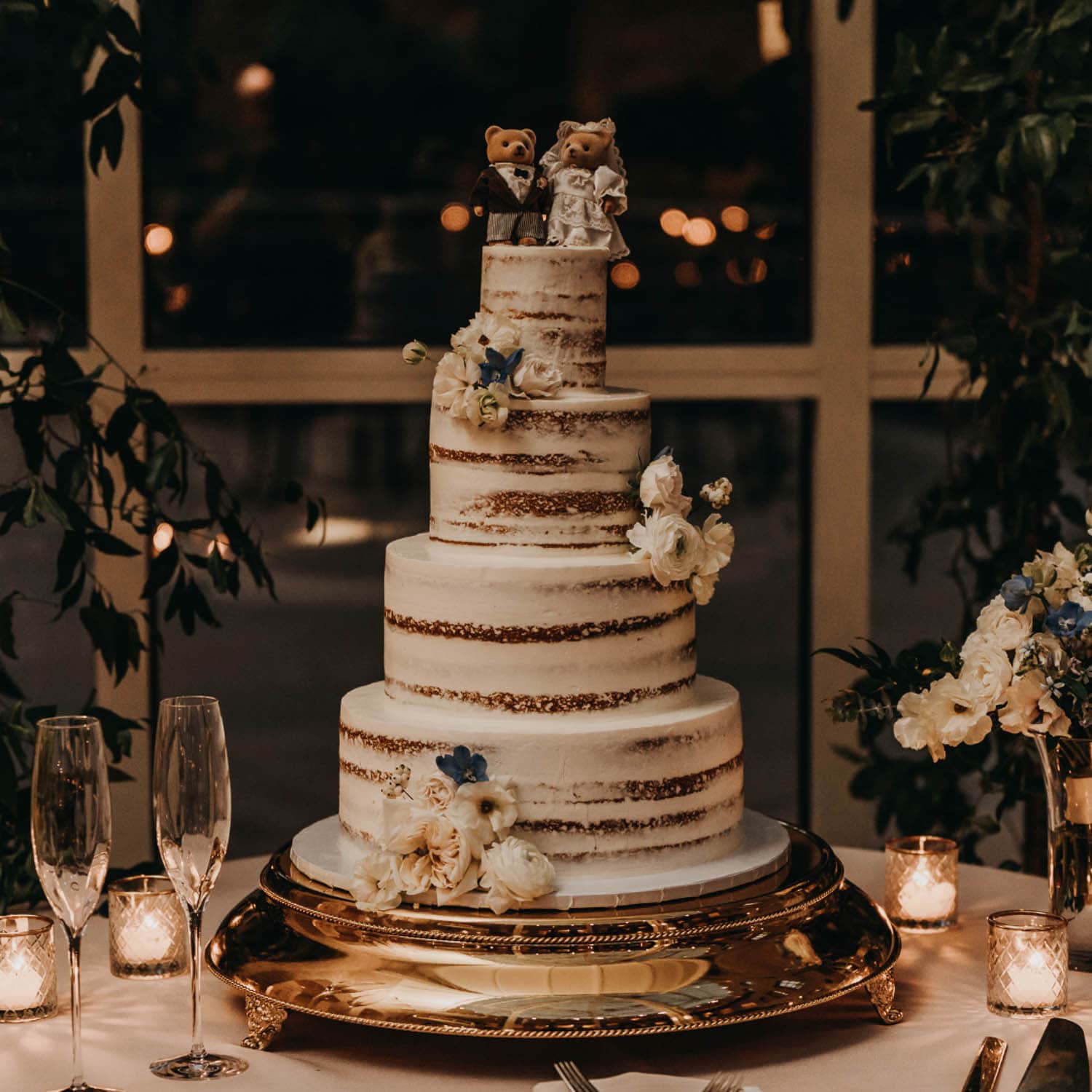 Four-tiered semi-naked wedding cake with white flowers and bear bride and groom toppers, on a gold stand with candles, flowers, and champagne flutes.