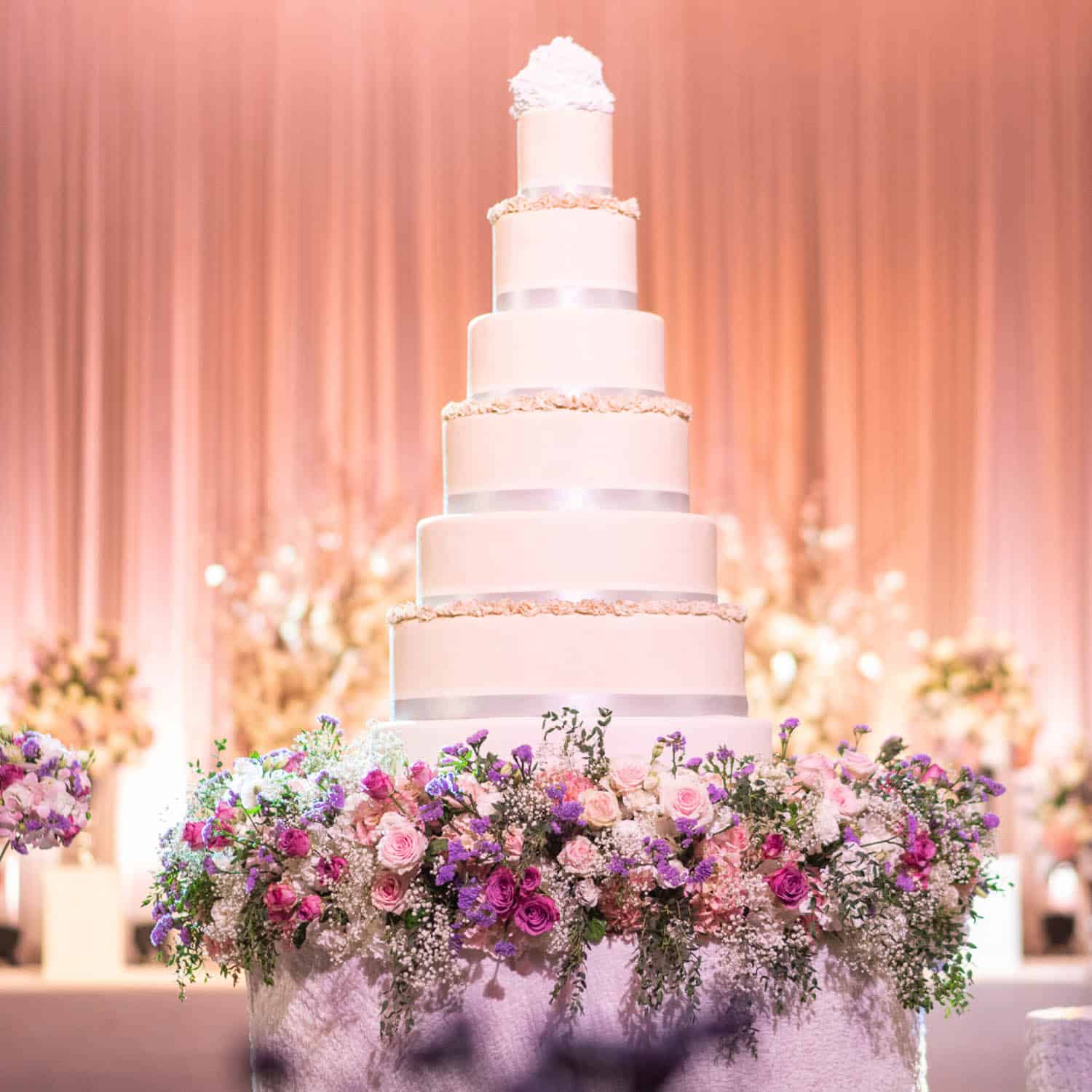A large, five-tiered white wedding cake with silver bands and floral decorations, surrounded by an arrangement of pink and purple flowers.