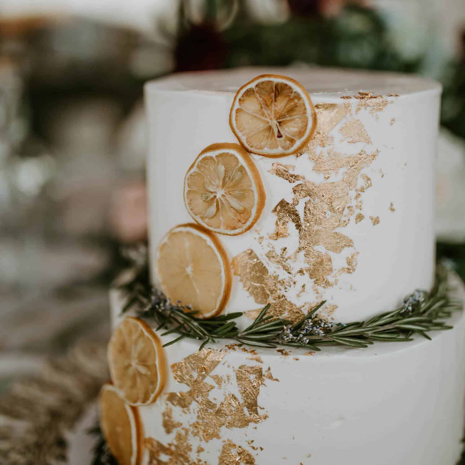 A white, two-tiered cake decorated with gold leaf, dried lemon slices, and a sprig of rosemary.