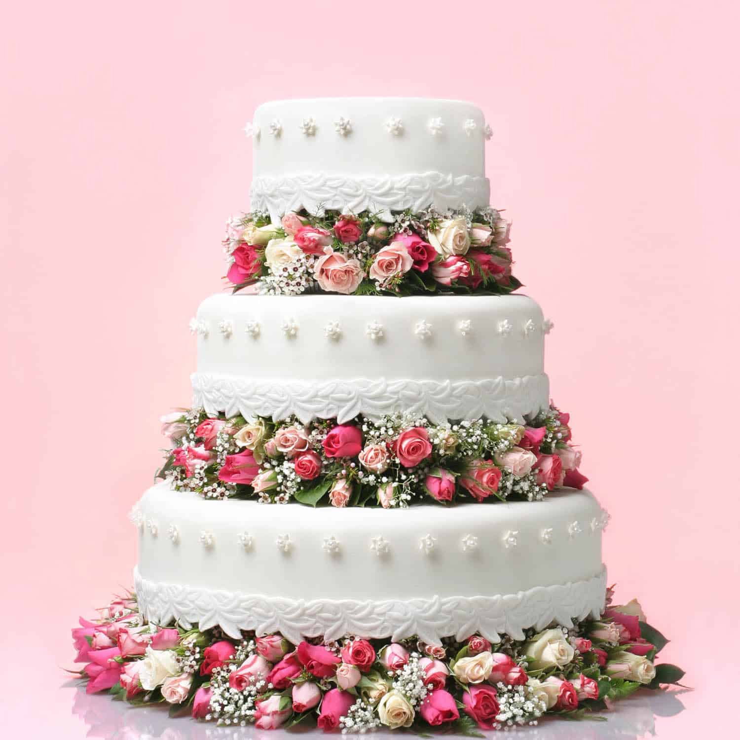 Three-tiered white wedding cake decorated with pink and white roses and baby's breath flowers, set against a pale pink background.