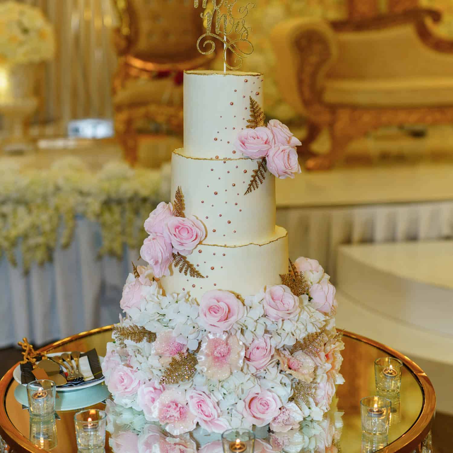 A three-tier white wedding cake decorated with pink roses, gold accents, and surrounded by white flowers, displayed on a glass table with small candles.