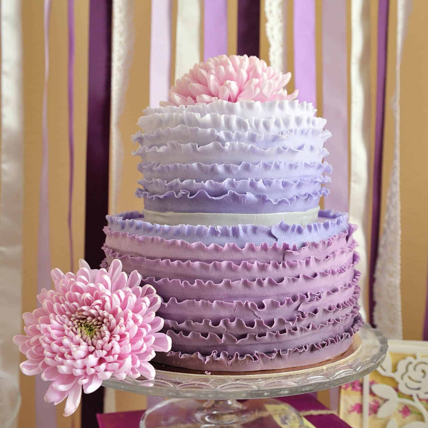 Two-tiered cake with purple ombré ruffled icing, large pink flowers on top and sides, set against a backdrop of vertical ribbon streamers.