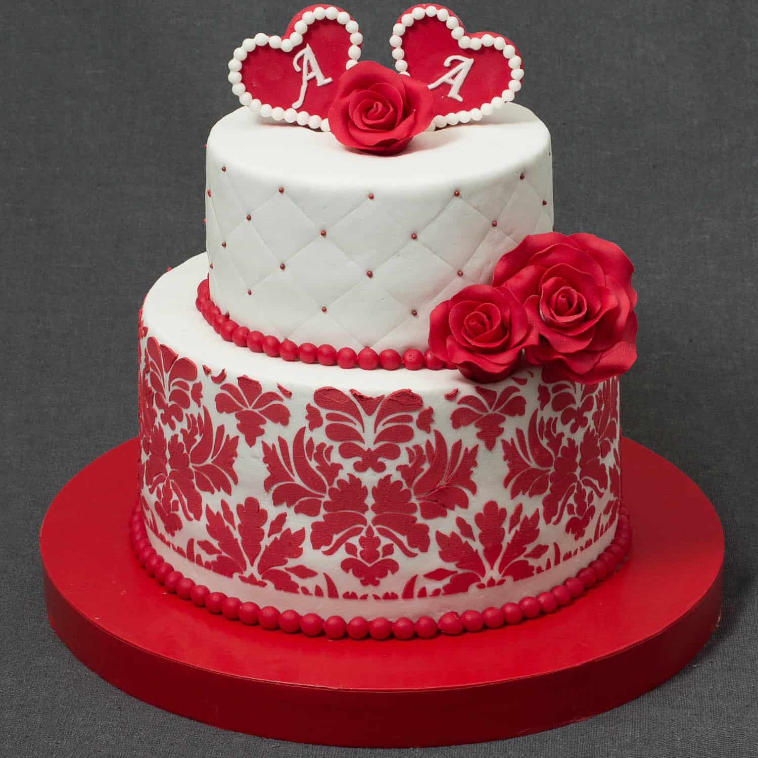 Two-tiered cake with red and white floral patterns, red roses, and two heart toppers with the letter "A" on top, displayed on a red base.