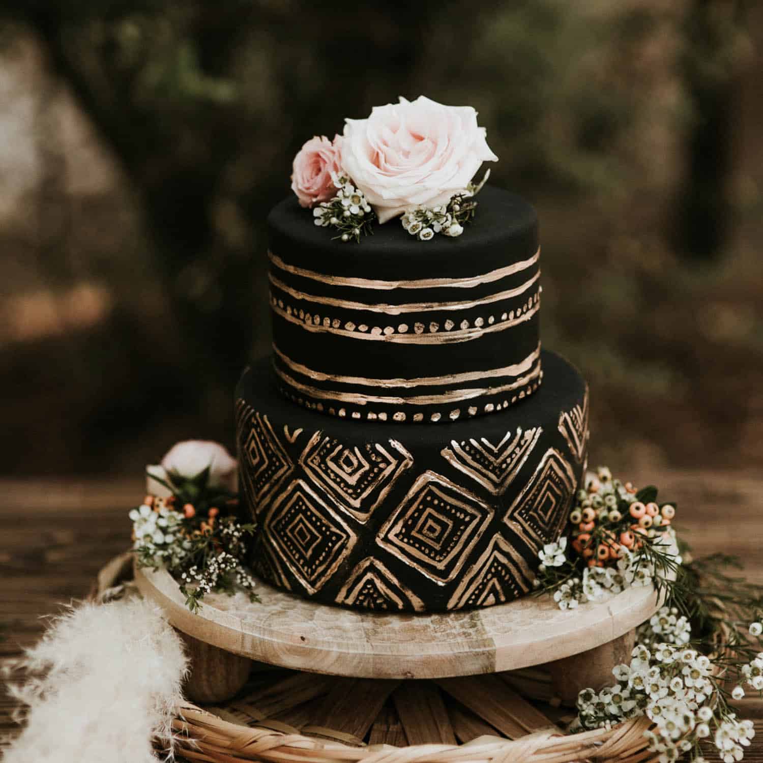 Two-tiered black cake with gold geometric and dotted patterns, topped with pink roses and greenery, displayed on a wooden stand with floral accents.