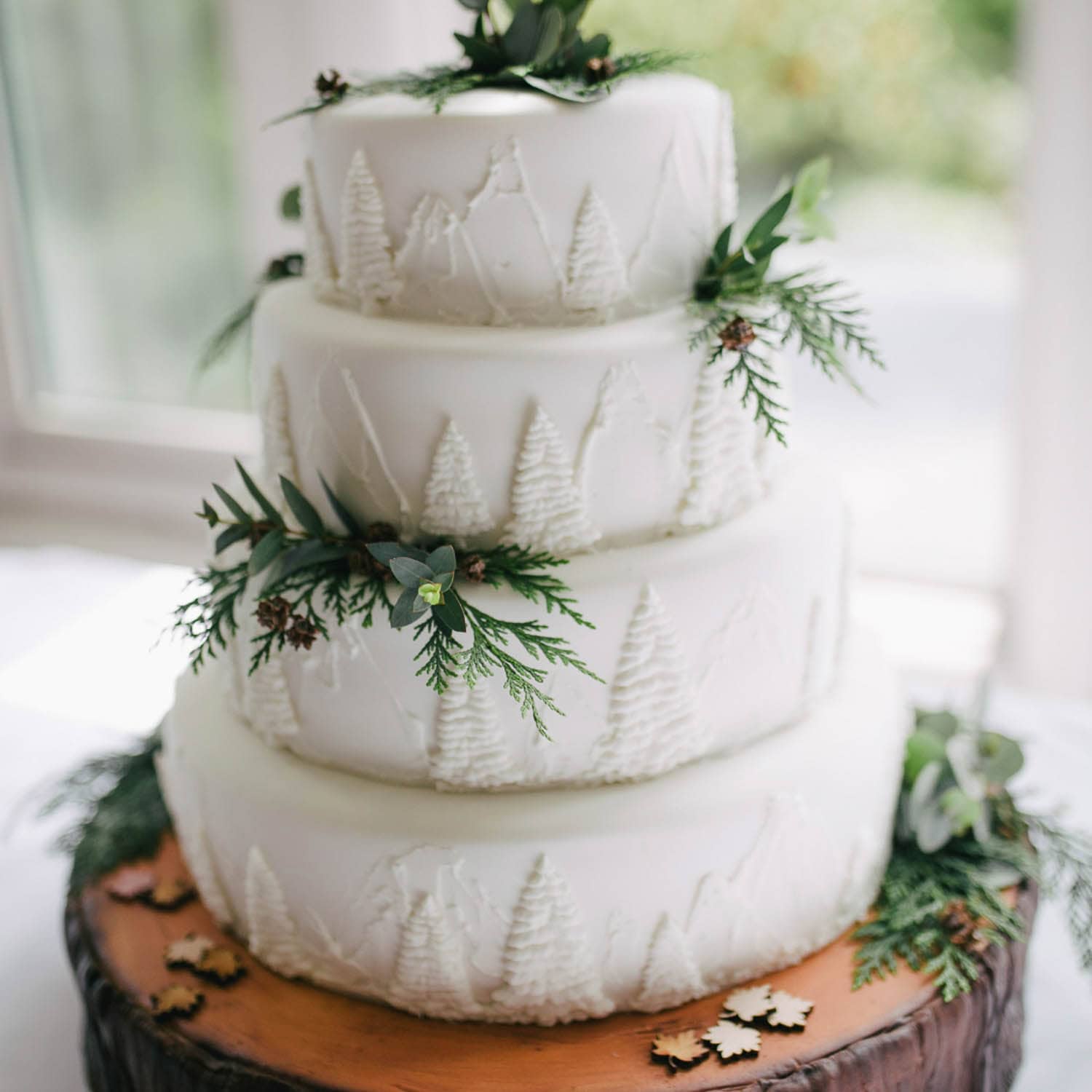 A three-tiered white cake decorated with piped trees, greenery, and small sprigs, displayed on a wooden base with a nature-inspired design.