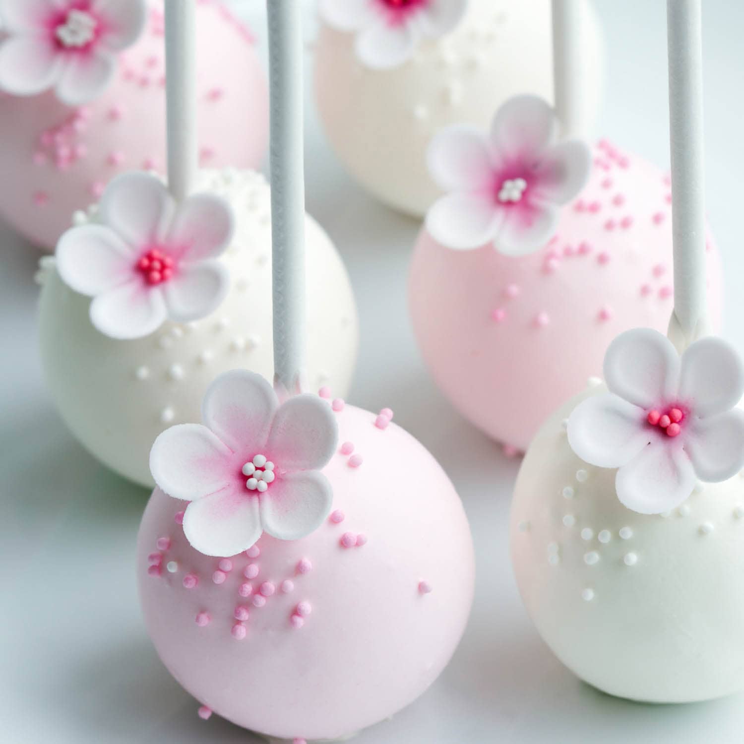 Pink and white cake pops decorated with small sugar flowers and sprinkles, arranged on a white surface.