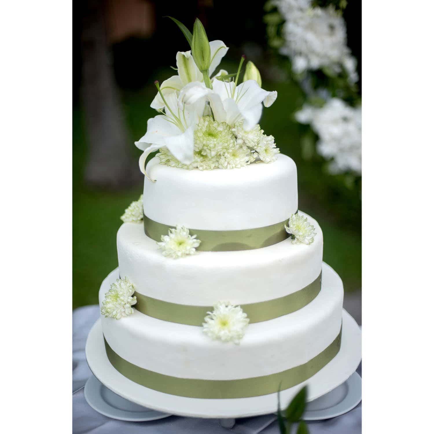 A three-tiered white cake decorated with green ribbons, white flowers, and topped with lilies, displayed outdoors.