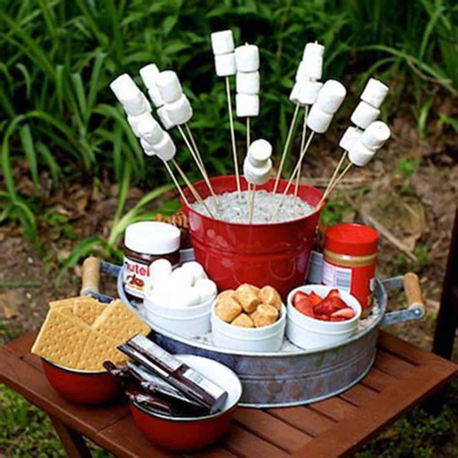 A tabletop s'mores station with marshmallows on skewers in a red bucket, surrounded by graham crackers, chocolate, strawberries, Nutella, and peanut butter.