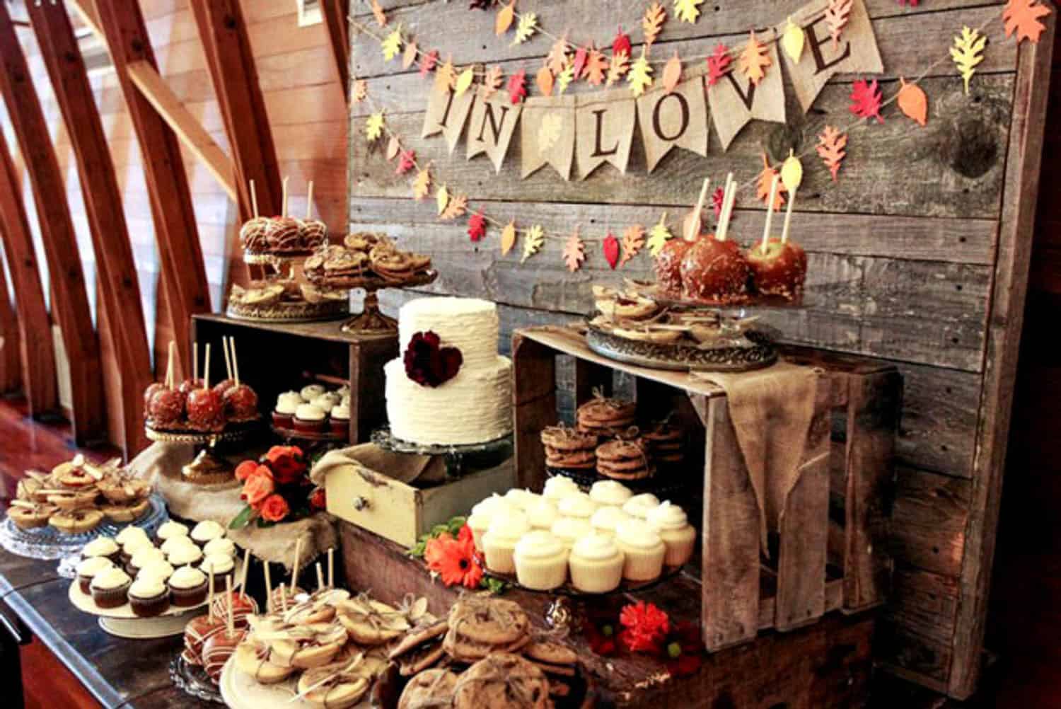 A rustic dessert table with cookies, cupcakes, caramel apples, a tiered cake, and fall decor, set against a wood backdrop with a bunting that reads "IN LOVE".
