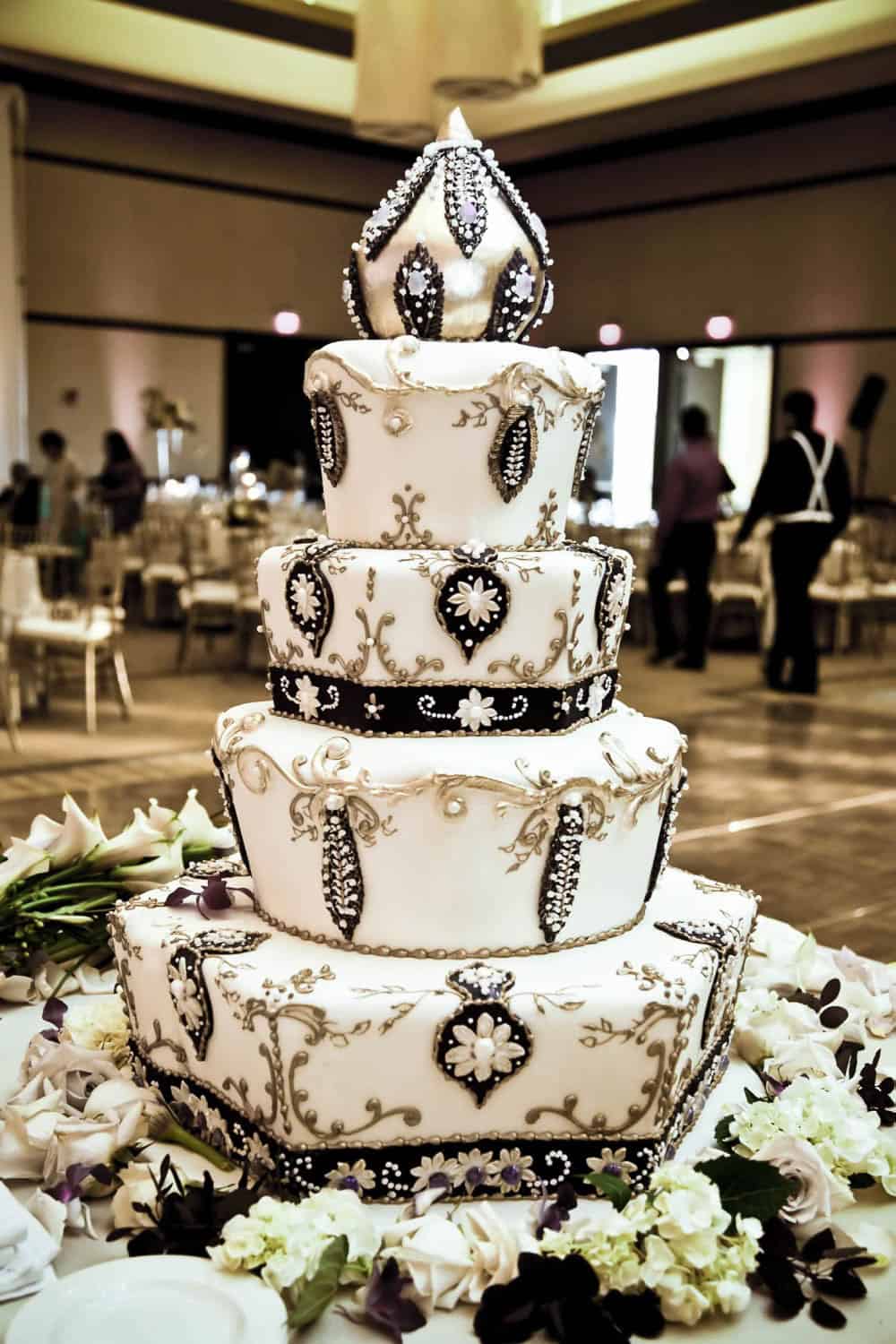 A four-tiered white wedding cake with ornate black and gold decorations, topped with a crown-like ornament, displayed on a table with flowers in a banquet hall.