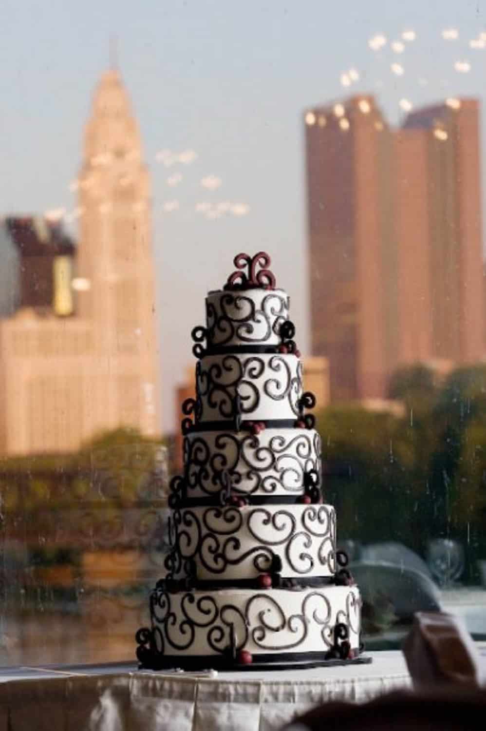 A five-tier white cake with black swirl decorations and red accents is displayed in front of a window overlooking a city skyline.